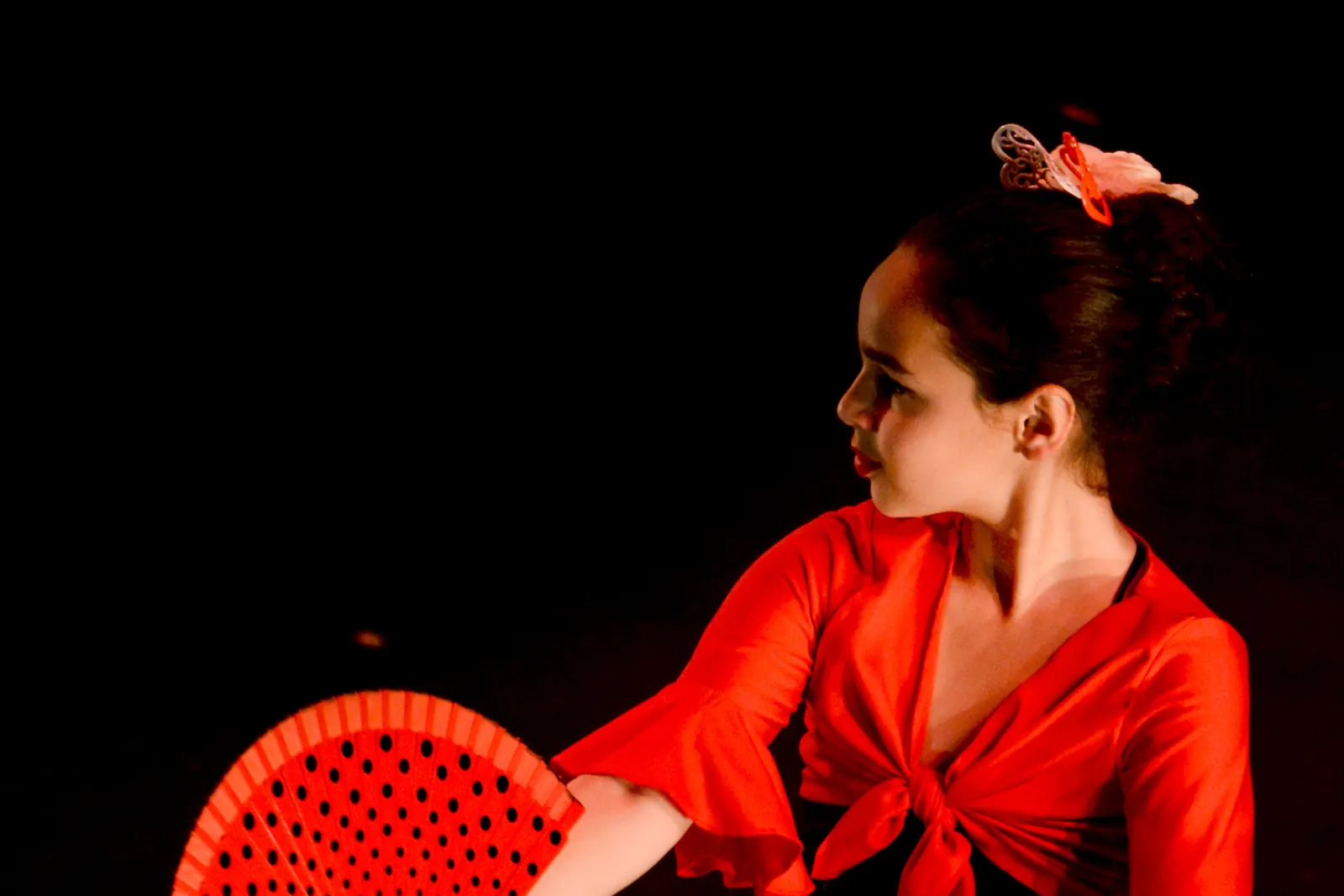 A dancer in a spotted red and black dress half hidden behind a red fan against a dark backdrop