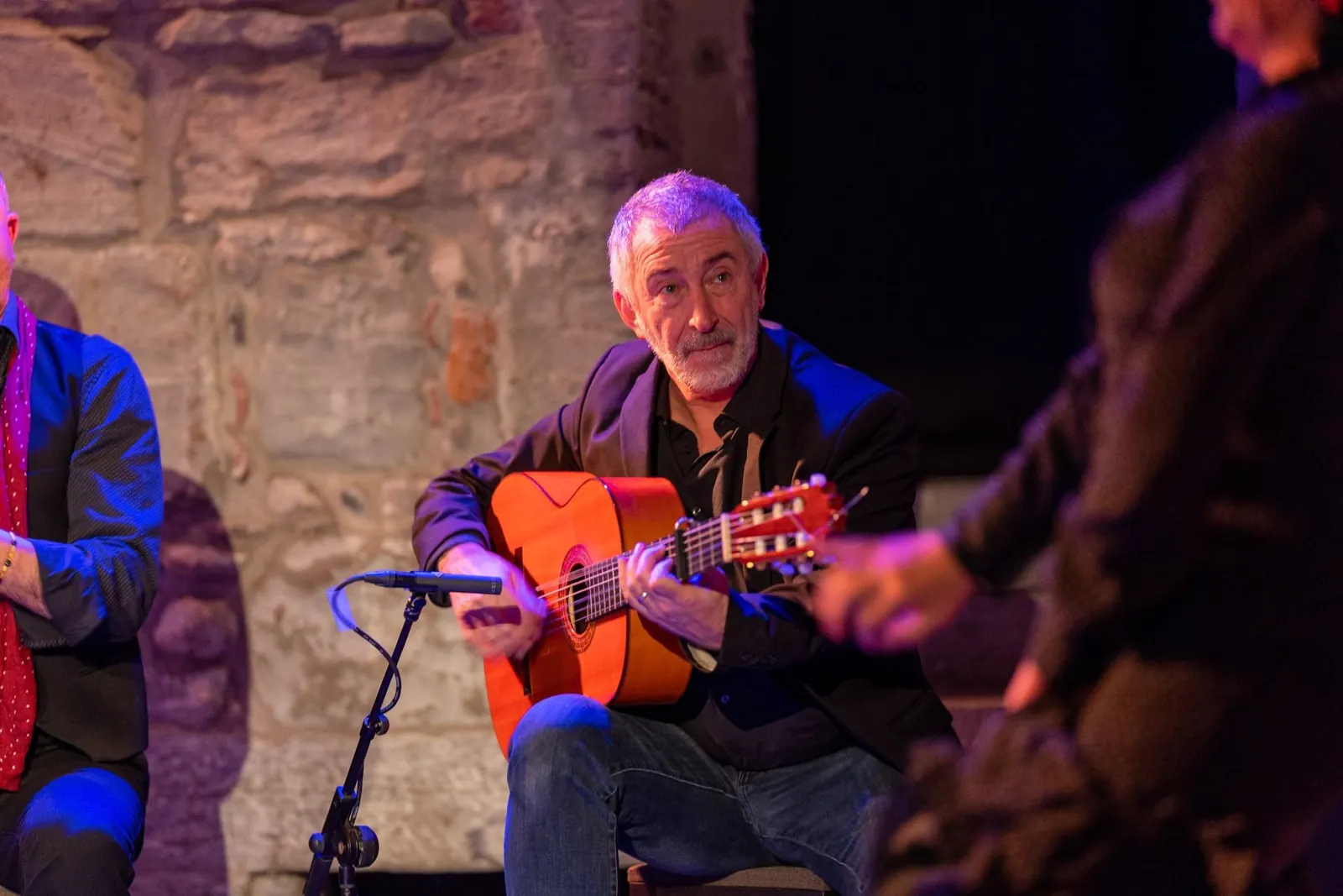 A flamenco guitarist in a dark jacket playing under stage lights beside a stone wall, with another performer clapping beside him