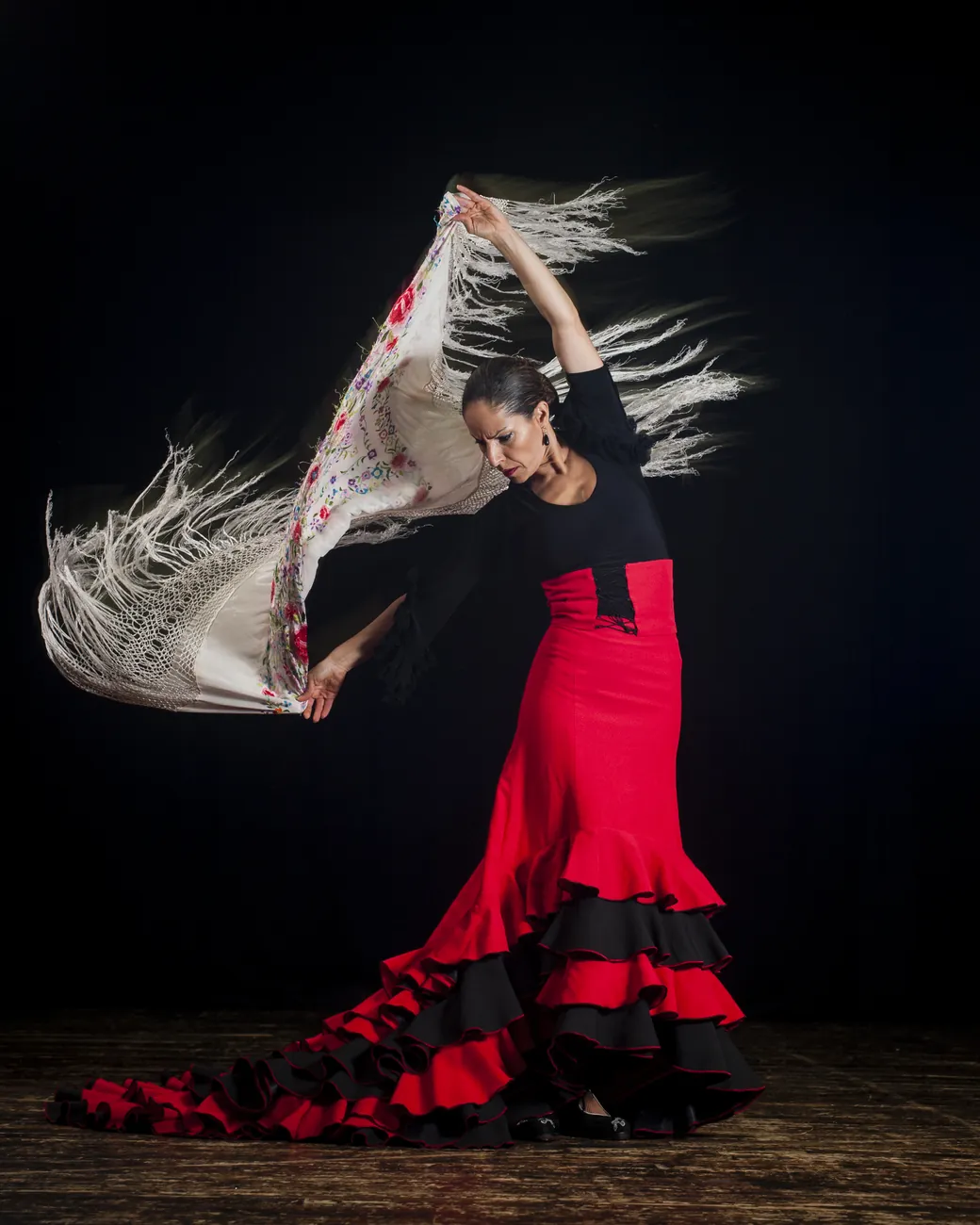 A flamenco dancer in a black and red dress swirling a fringed shawl against a black stage backdrop
